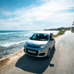 Χέρι γυναίκας που βγαίνει από το παράθυρο αυτοκινήτου, κρατώντας ψάθινο καπέλο, με θάλασσα και ιστιοπλοϊκό στο φόντο.