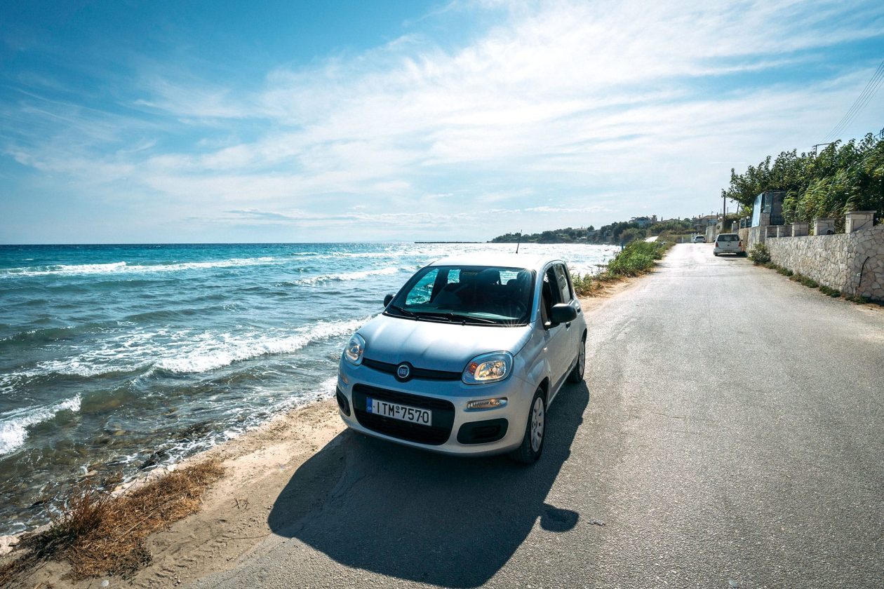Ασημένιο Fiat Panda παρκαρισμένο σε δρόμο δίπλα στη θάλασσα, με κύματα να σκάνε στην ακτή και γαλάζιο ουρανό με λευκά σύννεφα ψηλά.